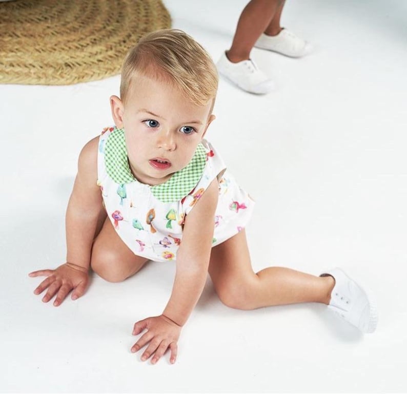 May include: A sleeveless white romper featuring a colorful bird print pattern and a green and white gingham collar. The garment has a button-down front closure and is worn by a toddler with light hair who is positioned on a white surface.