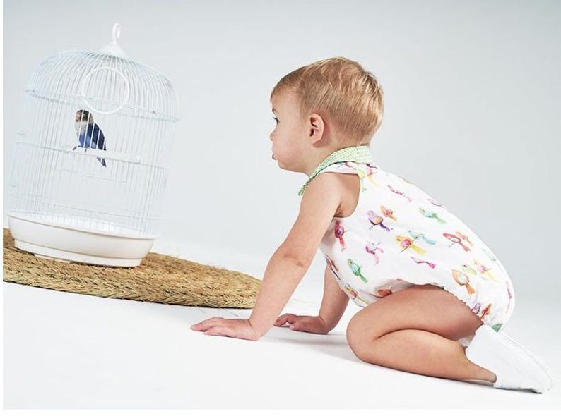 May include: A toddler wearing a white sleeveless romper with a colorful bird print and a green gingham collar, crawling toward a white wire birdcage containing a small blue and black lovebird perched on a white plastic rod inside the cage.