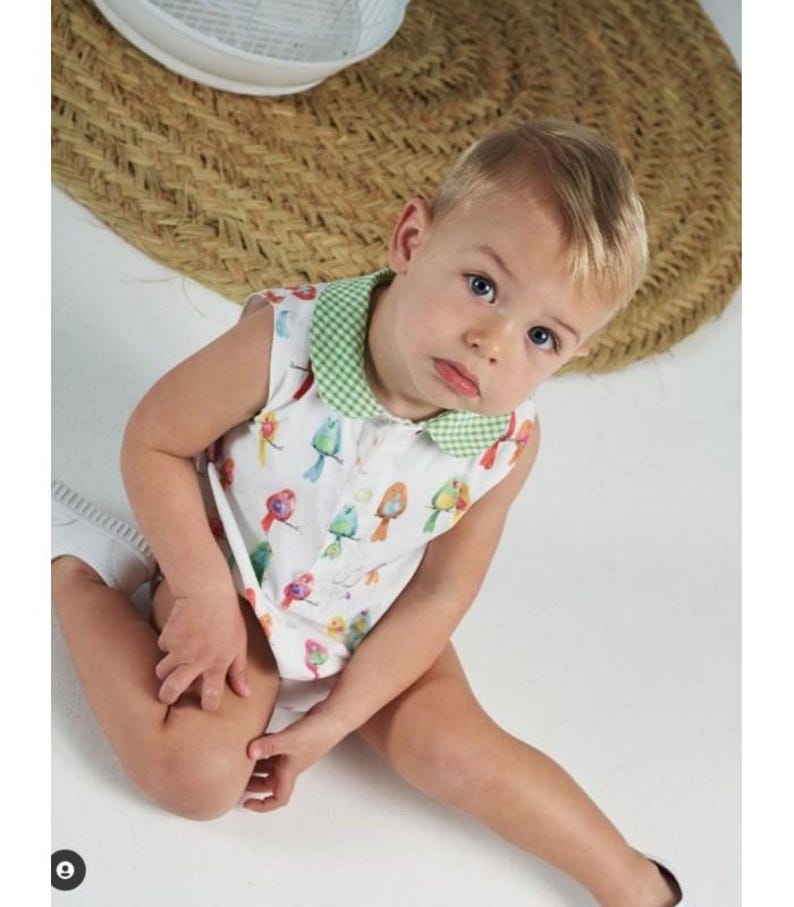 May include: A sleeveless white romper featuring a colorful bird pattern and a green and white gingham collar. The garment has a button-down front closure and is constructed from a lightweight fabric, providing a simple and functional design.
