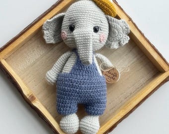 Può includere: Un pupazzo a forma di elefante lavorato all'uncinetto in grigio chiaro, che indossa una salopette blu e un piccolo basco giallo senape. L'elefante ha occhi a bottone neri, guance ricamate in rosa e un'etichetta rotonda con scritto Hand made.