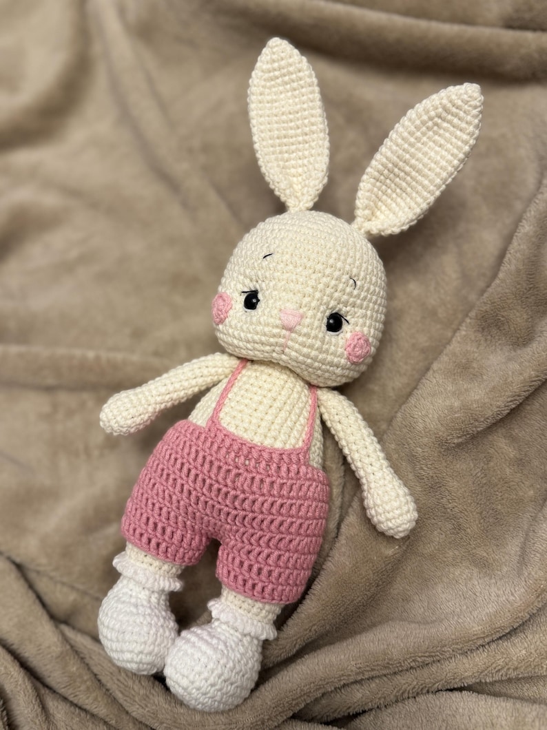 May include: A handmade crochet bunny doll with long ears, rosy cheeks, and black eyes. The bunny wears pink overalls with white suspenders and white ruffled socks. The doll is made of cream-colored yarn.