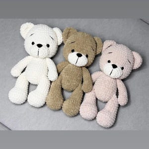 May include: Three handmade crochet teddy bears in cream, brown, and pink. Each bear has a black stitched nose and eyes, and a simple stitched mouth. The bears are lying on a grey surface.