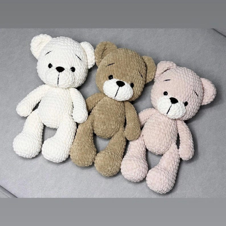 May include: Three handmade crochet teddy bears in cream, brown, and pink. Each bear has a black stitched nose and eyes, and a simple stitched mouth. The bears are lying on a grey surface.