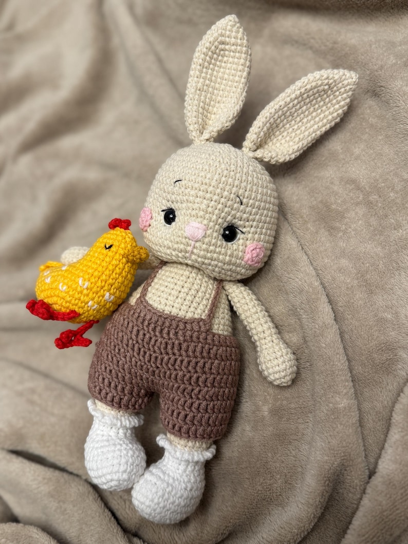 May include: A handmade crochet bunny doll with a small yellow chick. The bunny has long ears, pink cheeks, and is wearing brown overalls and white socks. The chick has red accents. The toys are made of yarn.