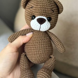 May include: A brown crocheted teddy bear with black eyes, a white nose, and a white muzzle. The bear is sitting on a light brown surface.