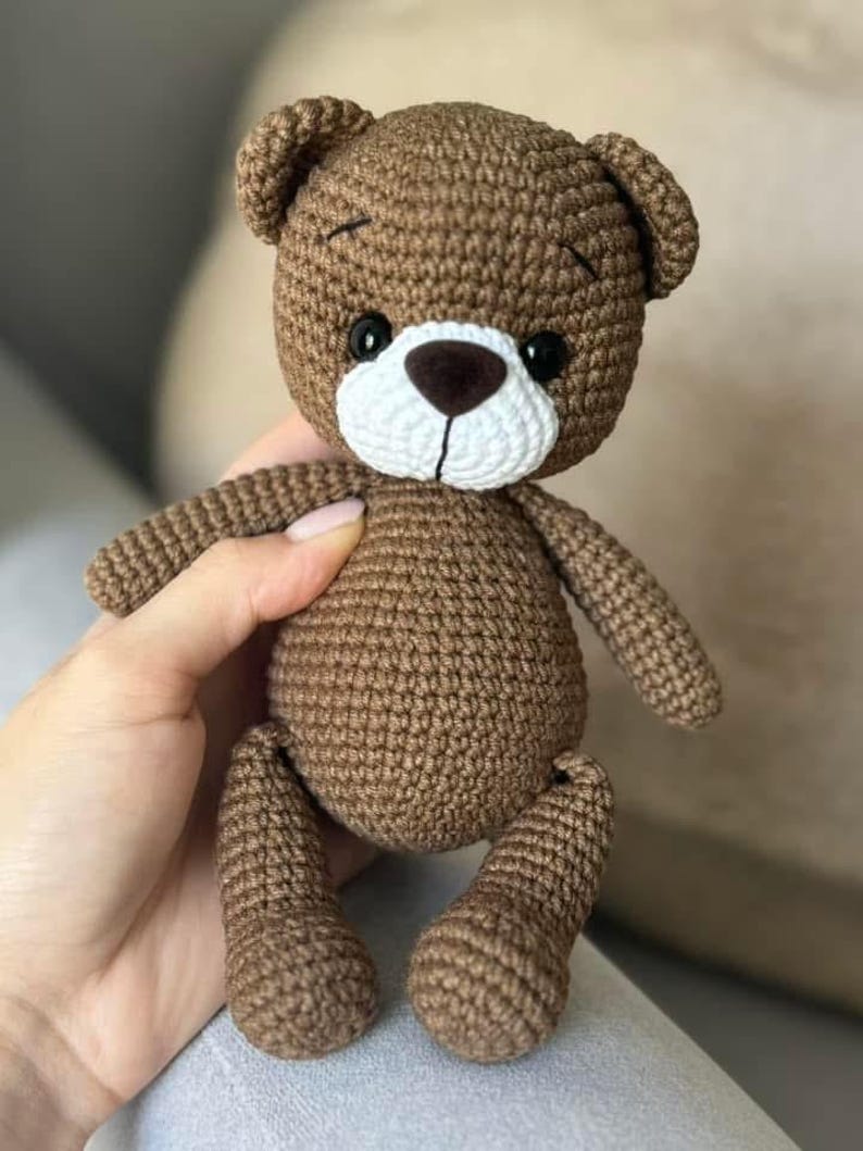 May include: A brown crocheted teddy bear with black eyes, a white nose, and a white muzzle. The bear is sitting on a light brown surface.