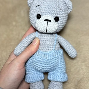May include: A gray crocheted teddy bear wearing blue overalls. The bear has black eyes and a white nose.