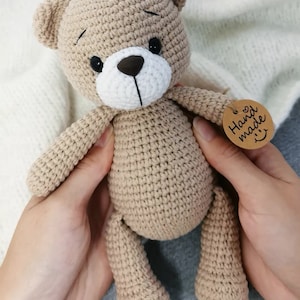 May include: A crocheted brown teddy bear with black eyes and a white nose. The bear is being held by two hands and has a brown tag that says "Hand made" with a heart and a smiley face.