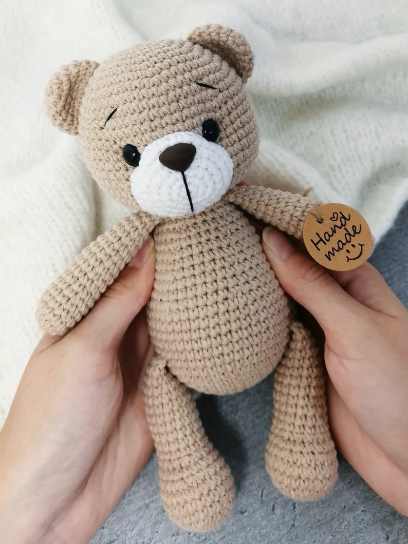 May include: A crocheted brown teddy bear with black eyes and a white nose. The bear is being held by two hands and has a brown tag that says "Hand made" with a heart and a smiley face.