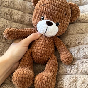 May include: A handmade brown teddy bear with a white muzzle and black eyes. The bear is made of a soft, textured material and has a classic teddy bear shape with rounded features. The bear is being held by a person.