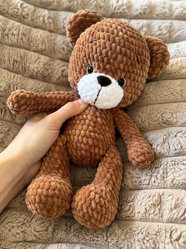 May include: A handmade brown teddy bear with a white muzzle and black eyes. The bear is made of a soft, textured material and has a classic teddy bear shape with rounded features. The bear is being held by a person.