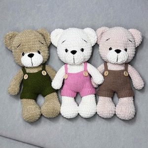 May include: Three crocheted teddy bears. One is beige with green dungarees, the second is white with pink dungarees, and the third is pale pink with brown dungarees. Each bear has black eyes and a black stitched nose.