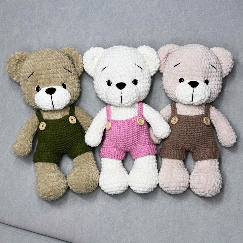 May include: Three crocheted teddy bears. One is beige with green dungarees, the second is white with pink dungarees, and the third is pale pink with brown dungarees. Each bear has black eyes and a black stitched nose.