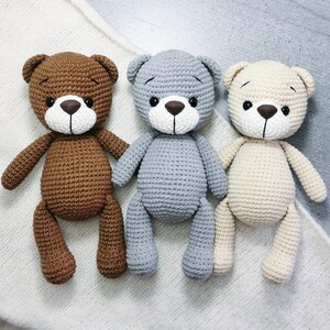 May include: Three crocheted teddy bears, one brown, one gray, and one beige, sitting on a white fuzzy surface.