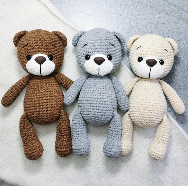 May include: Three crocheted teddy bears, one brown, one gray, and one beige, sitting on a white fuzzy surface.