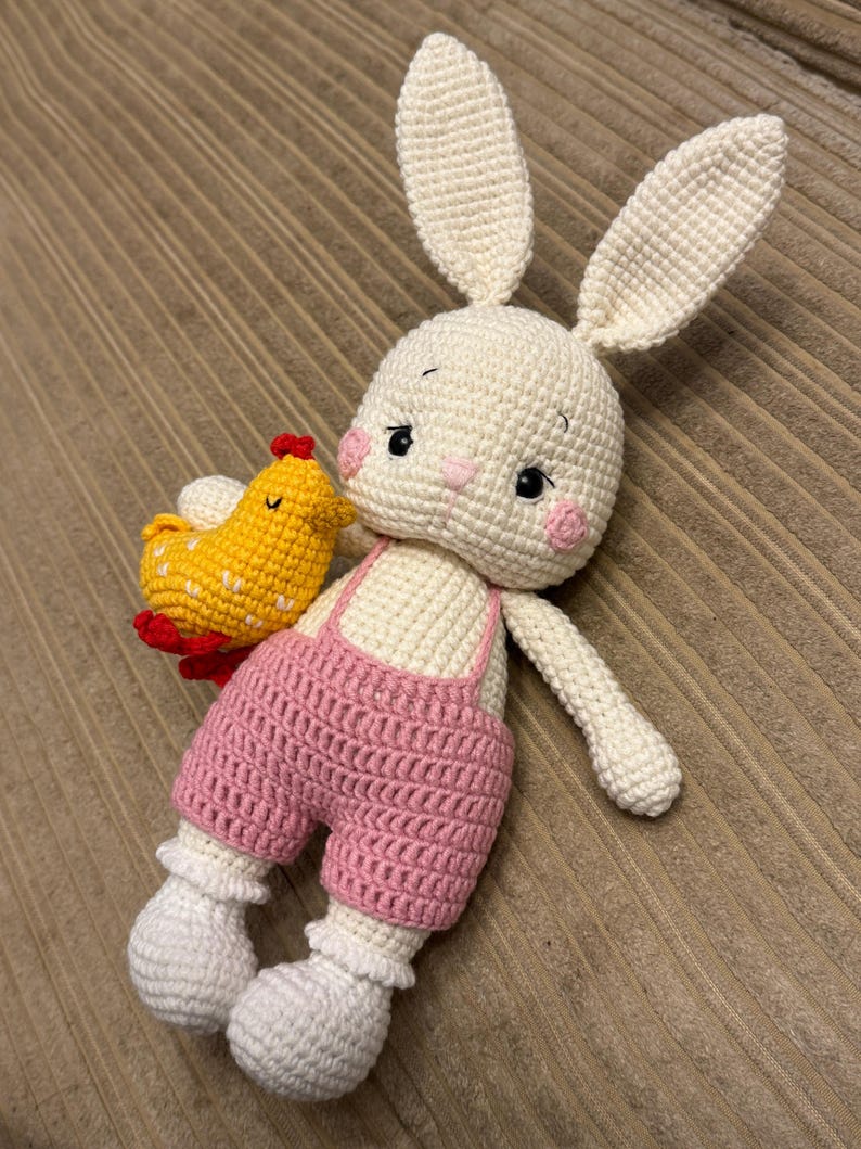 May include: A handmade crochet bunny doll with a yellow chick. The bunny is white with pink cheeks and overalls. The chick is yellow with red accents. The doll is lying on a brown surface.