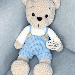May include: A soft, beige teddy bear wearing a white shirt and light blue dungarees with wooden button details. The bear has a brown nose and black eyes. A tag that says "Hand made" is attached.
