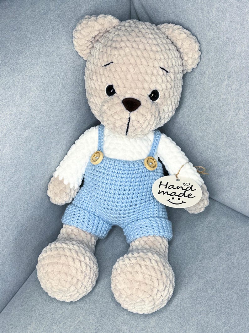 May include: A soft, beige teddy bear wearing a white shirt and light blue dungarees with wooden button details. The bear has a brown nose and black eyes. A tag that says "Hand made" is attached.