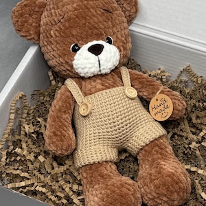 May include: A brown crocheted teddy bear wearing beige overalls with wooden buttons. The bear has a white muzzle and a small tag that says "Hand made". The bear is lying in a box filled with brown paper.