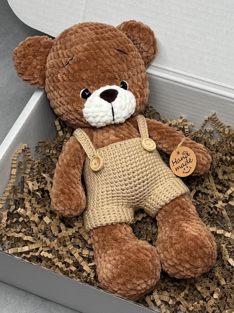 May include: A brown crocheted teddy bear wearing beige overalls with wooden buttons. The bear has a white muzzle and a small tag that says "Hand made". The bear is lying in a box filled with brown paper.