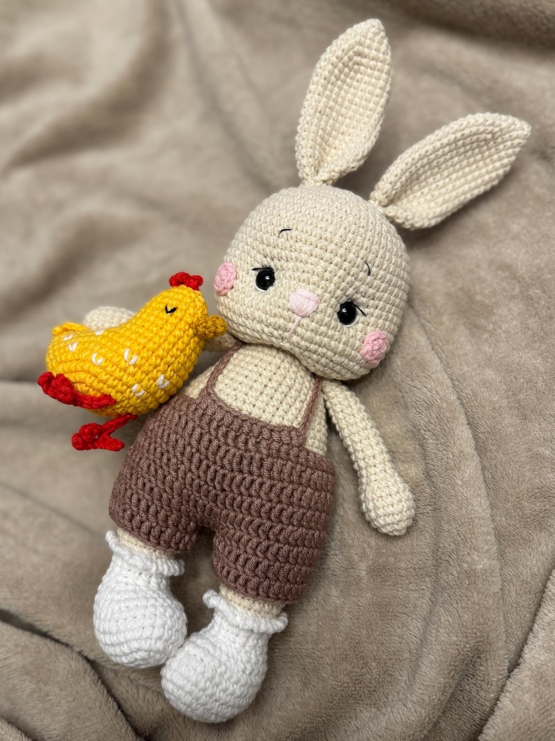 May include: A handmade crochet bunny doll with a yellow chicken. The bunny has long ears, pink cheeks, and brown overalls. The chicken is yellow with red accents. The doll is made of soft yarn and is a cute, cuddly toy.