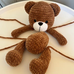 May include: A brown crocheted teddy bear with a white muzzle and black eyes. The bear's arms and legs are detached, with long yarn strands extending from each limb. The bear is made of a soft, textured yarn.