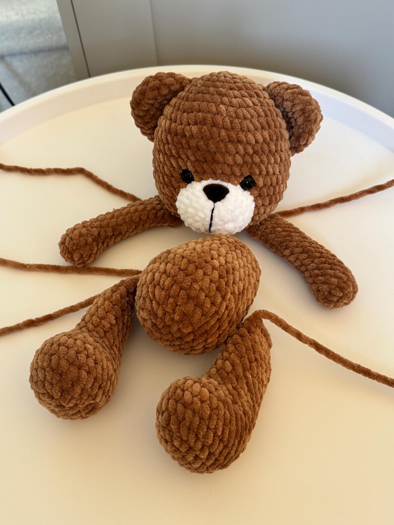 May include: A brown crocheted teddy bear with a white muzzle and black eyes. The bear's arms and legs are detached, with long yarn strands extending from each limb. The bear is made of a soft, textured yarn.