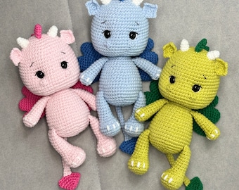 May include: Three crocheted dragon toys. One is pink, one is light blue, and one is green. Each dragon has a small, embroidered face and wings.