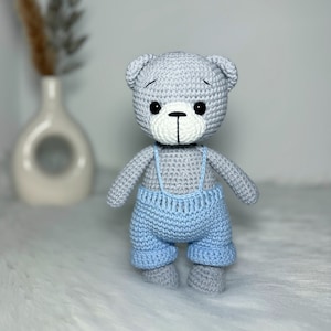 May include: A gray crocheted teddy bear wearing blue overalls. The bear has black eyes and a white nose.