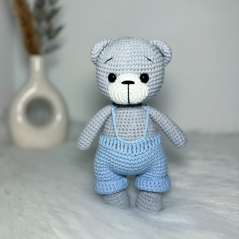 May include: A gray crocheted teddy bear wearing blue overalls. The bear has black eyes and a white nose.