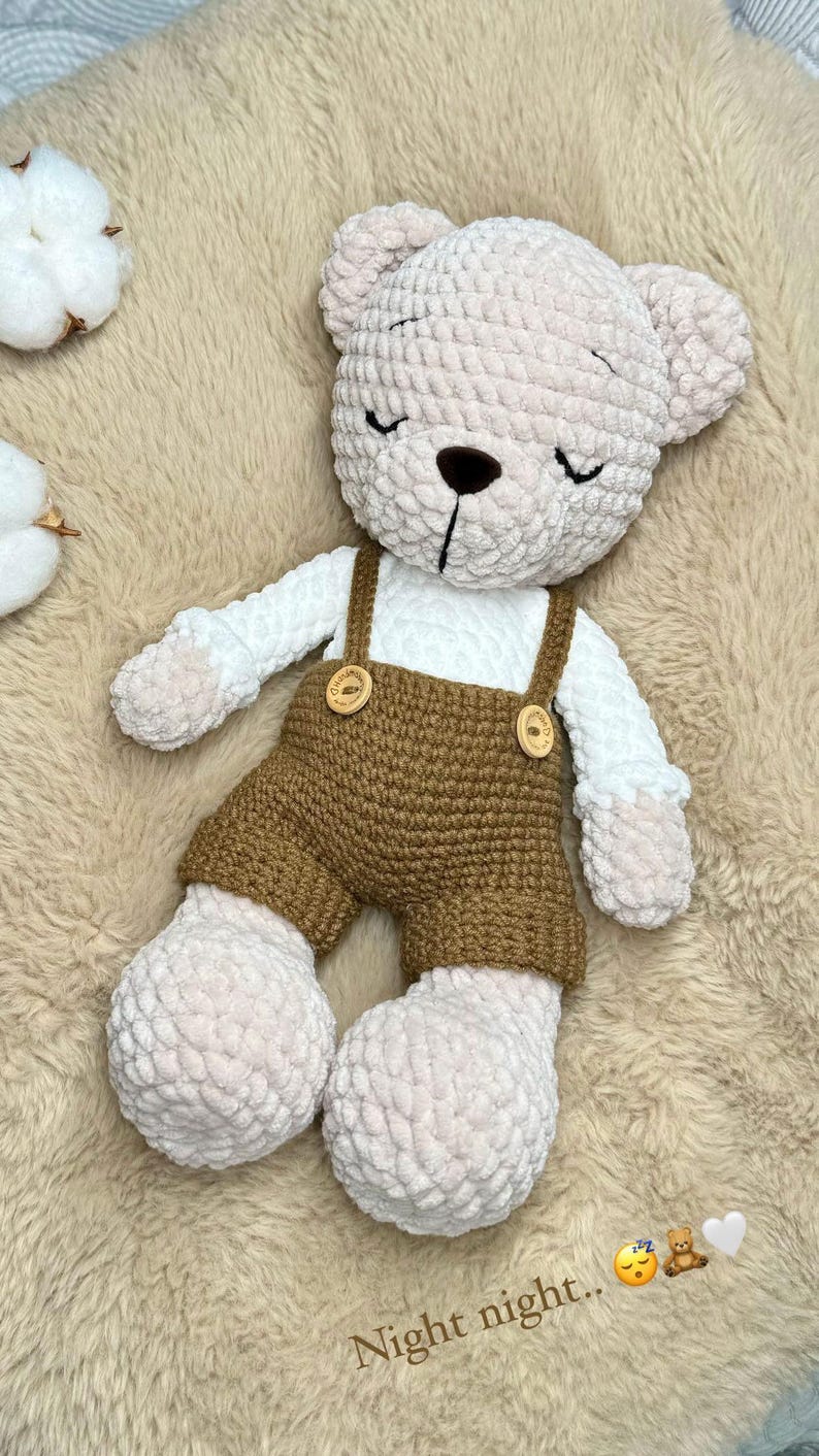 May include: A crocheted teddy bear with a soft, plush texture. The bear is dressed in a white shirt and brown overalls with wooden buttons. The bear's eyes are closed, and the text "Night night.." is visible.