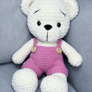 May include: A handmade white crochet teddy bear wearing pink dungarees with wooden button fastenings. The bear has black eyes and a black stitched nose and mouth. The bear is sitting on a grey surface.