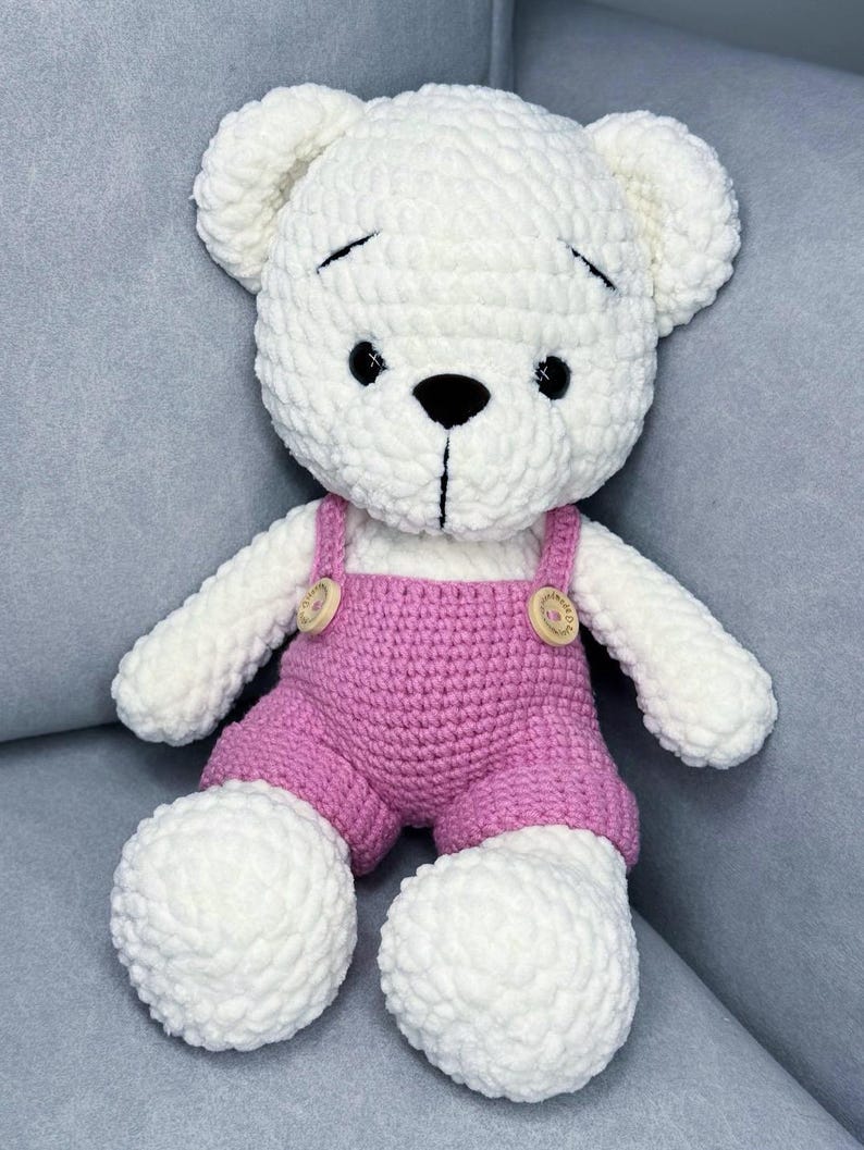 May include: A handmade white crochet teddy bear wearing pink dungarees with wooden button fastenings. The bear has black eyes and a black stitched nose and mouth. The bear is sitting on a grey surface.