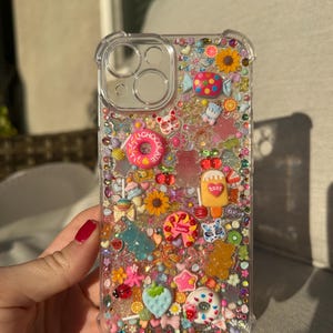 May include: A clear phone case with a colorful, glittery design featuring various sweets, including a donut, candy canes, lollipops, and gummy bears. The case is filled with glitter and small, colorful beads.