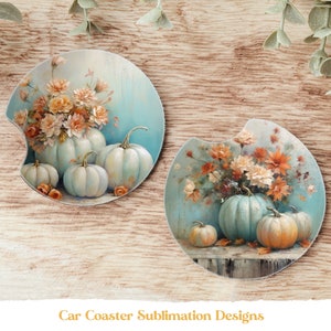 Op de afbeelding: Twee auto-onderzetters met een herfstthema. Elke onderzetter heeft een blauwe achtergrond met een boeket oranje en witte bloemen en een groep witte pompoenen. De pompoenen zijn lichtblauw geverfd.