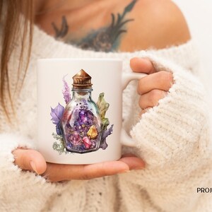 Peut inclure: Mug en c&eacute;ramique blanche avec une illustration &agrave; l'aquarelle d'une bouteille en verre remplie de cristaux color&eacute;s et d'un bouchon en li&egrave;ge. La bouteille est entour&eacute;e de feuillage violet et vert. Le mug est tenu par une personne portant un pull blanc.