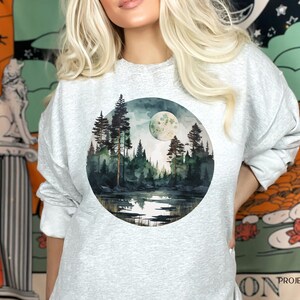May include: A gray sweatshirt with a circular design featuring a watercolor painting of a moonlit forest scene with a lake reflecting the moon.
