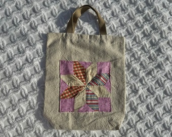 May include: A beige tote bag with a brown handle. The bag features a square patchwork design with a starburst pattern in shades of red, blue, and beige. The bag is made of a textured fabric.