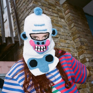 May include: A white crocheted balaclava-style hat with a blue pom-pom, blue ears, and a pink and blue face design. The hat covers the head and neck. The person is wearing a striped blue, red, and green shirt.