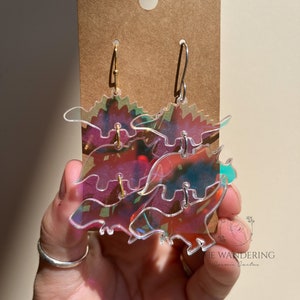 Peut inclure: Une paire de boucles d'oreilles en acrylique irisé en forme de dinosaures. Les boucles d'oreilles sont sur une carte brune avec le texte "The Wandering Cosmic Cactus".