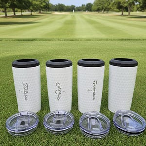 May include: Four white golf ball-textured tumblers with black rims and clear lids are displayed on a green golf course. Each tumbler has a different golf brand logo: Titleist, Callaway, and TaylorMade. The background shows a golf course.