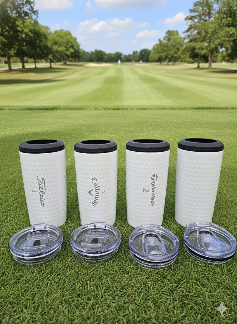 May include: Four white golf ball-textured tumblers with black rims and clear lids are displayed on a green golf course. Each tumbler has a different golf brand logo: Titleist, Callaway, and TaylorMade. The background shows a golf course.
