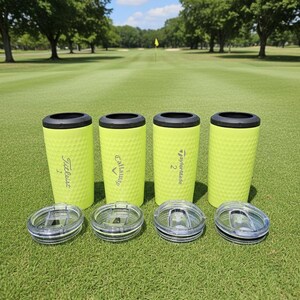 May include: Four neon green golf ball textured tumblers with black rims and clear lids are displayed on a golf course. The tumblers have the logos of golf brands Titleist, Callaway, and TaylorMade. The background shows a green golf course.