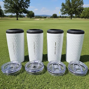 May include: Four white travel mugs with black rims and clear lids sit on green grass. The mugs have a dimpled surface, similar to golf balls, and feature the text "PRO V1X", "CHROME SOFT", and "TP5". The background shows a golf course under a blue sky.