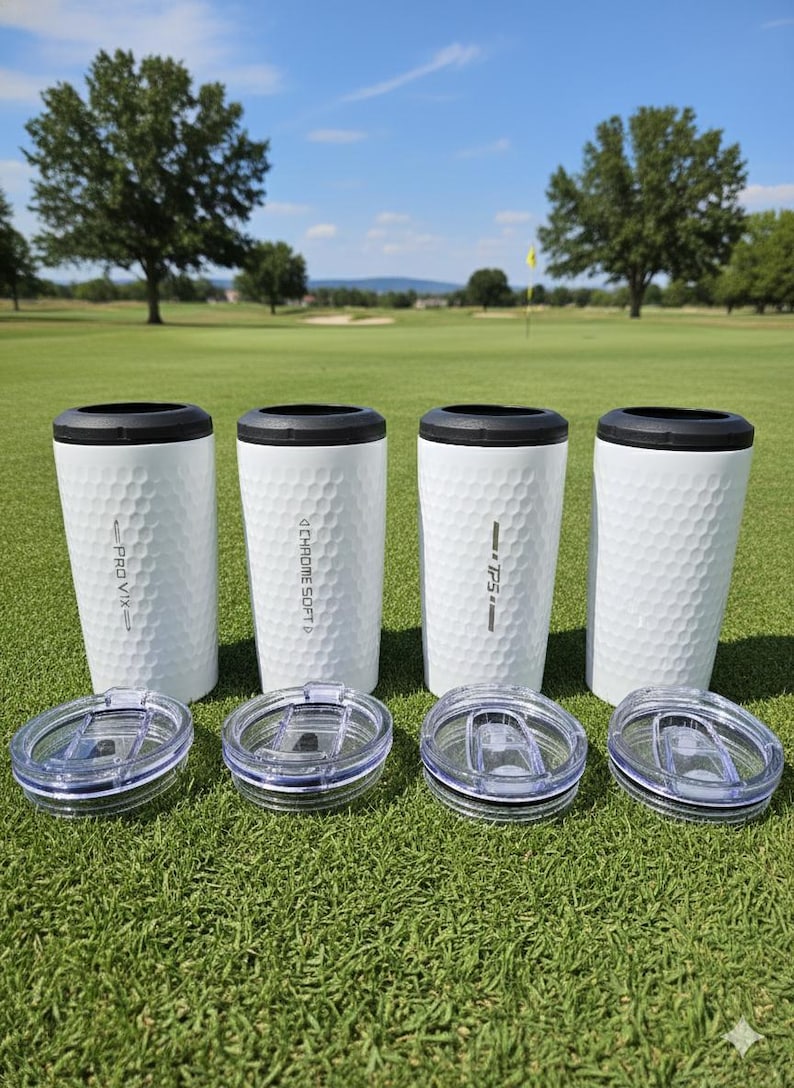 May include: Four white travel mugs with black rims and clear lids sit on green grass. The mugs have a dimpled surface, similar to golf balls, and feature the text "PRO V1X", "CHROME SOFT", and "TP5". The background shows a golf course under a blue sky.