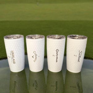 May include: Four white golf ball-textured tumblers with silver lids. Each tumbler features a different golf brand name: Kirkland Signature, Callaway, TaylorMade, and Titleist. The tumblers are displayed on a green surface.