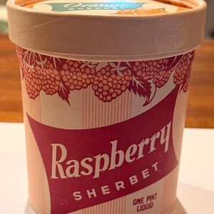 May include: A cylindrical paper pint container of Raspberry Sherbet with a light pink and white striped background. The label features a red banner with the text Raspberry Sherbet and One Pint Liquid. The lid shows Orange Sherbet text.