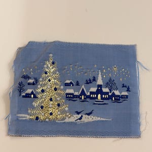 May include: A rectangular piece of light blue fabric featuring an embroidered winter village scene. The design includes a large gold and blue Christmas tree, small houses with white roofs, a church, and gold stars against a light blue background.