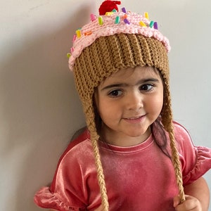 May include: A child wearing a pink and brown crocheted cupcake hat with colourful sprinkles and a red cherry on top. The hat has long brown crocheted ties.