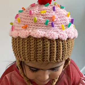 May include: A pink and brown crocheted cupcake hat with a red cherry on top and colourful sprinkles. The hat has ear flaps and is being worn by a child.
